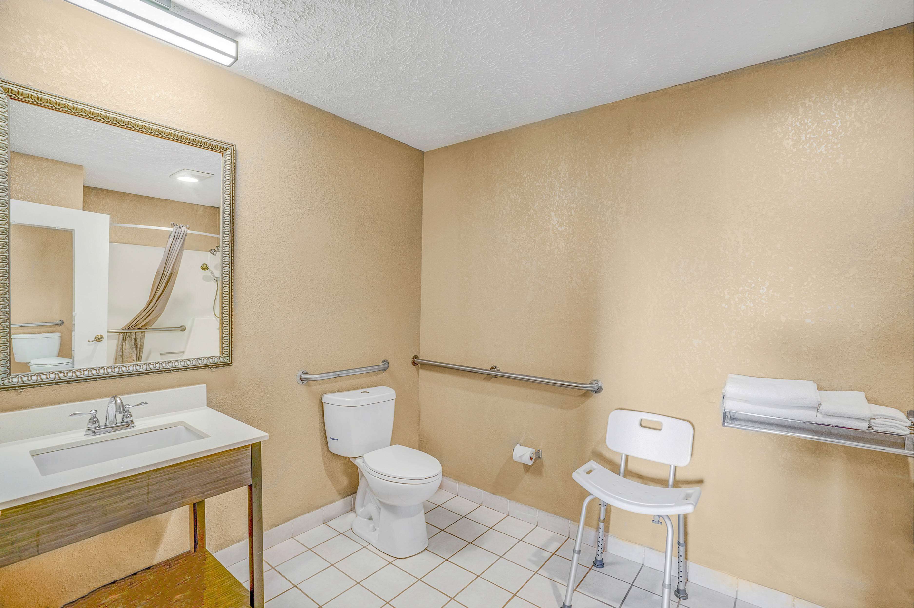Un baño con lavamanos, inodoro y ducha blancos. Hay un espejo con marco dorado, un perchero y una silla.