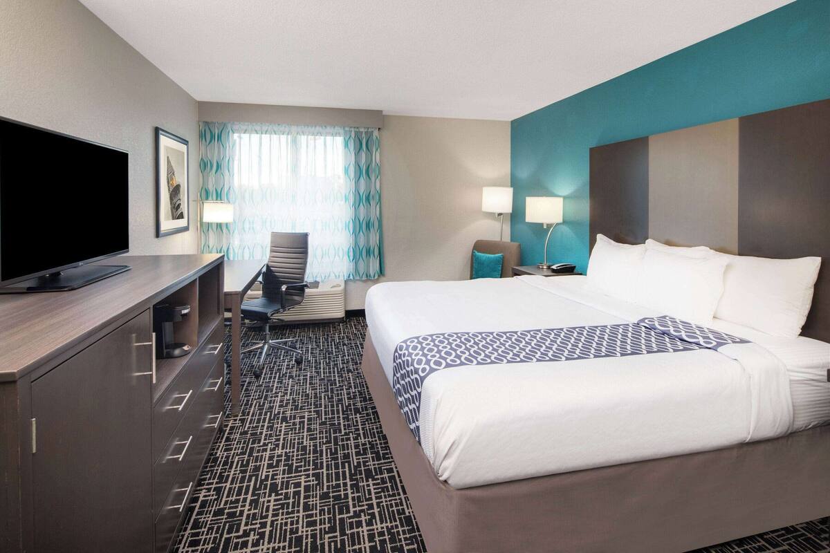 Une chambre d’hôtel comprenant un lit, un bureau avec un téléviseur à écran plat, une chaise, une lampe et une fenêtre avec des rideaux.