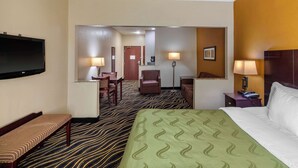 Ein Hotelzimmer mit einem großen Bett, einem an der Wand befestigten Fernseher, einem Schreibtisch mit Stuhl und einem kleinen Tisch mit Lampe.