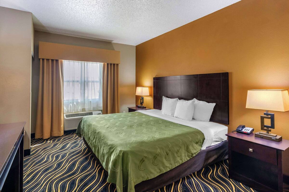 A hotel room with a large bed, two bedside tables with lamps, a window with curtains, and a patterned carpet.