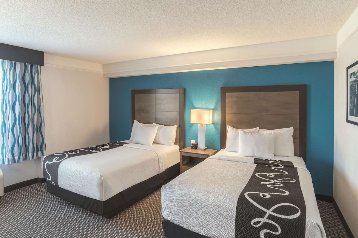 A hotel room with two beds, a nightstand with a lamp, and a blue accent wall.