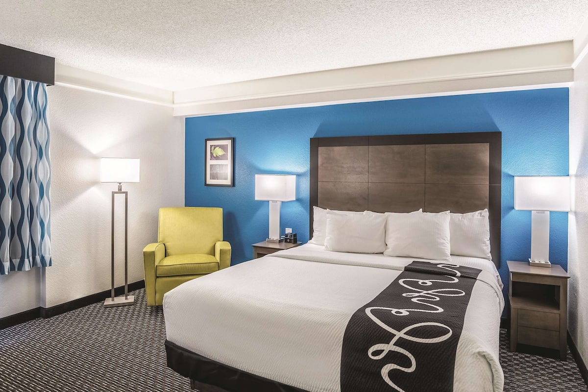 A hotel room with a large bed, a yellow armchair, two bedside lamps, and a framed picture on the wall.