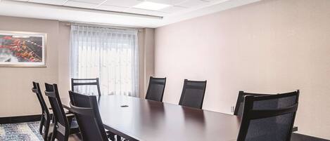 A conference room with a long table, six chairs, a window with sheer curtains, and a framed picture on the wall.