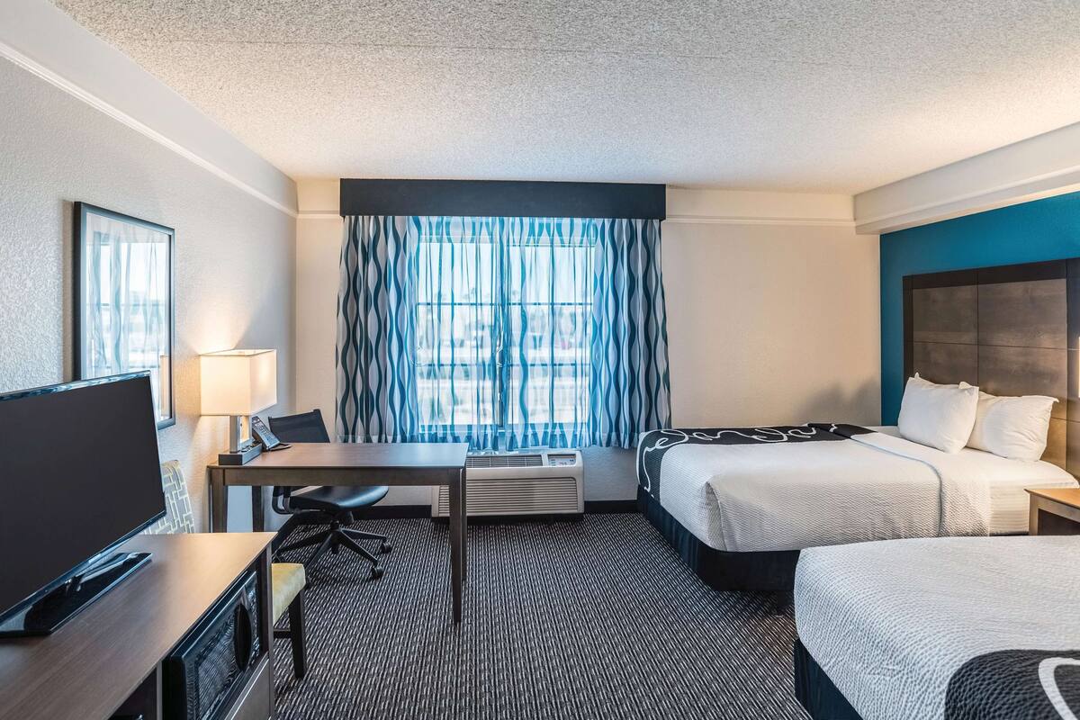 A hotel room with a bed, a desk with a chair, a television, and a window with curtains.