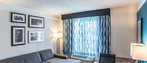 Habitación de hotel con sofá gris, una mesita, un escritorio con una silla, una lámpara de pie y una ventana con cortinas estampadas.