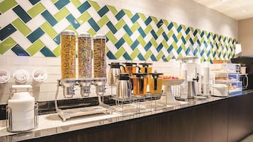 A breakfast buffet area with various food items and beverages, including cereal, fruits, and coffee machines.