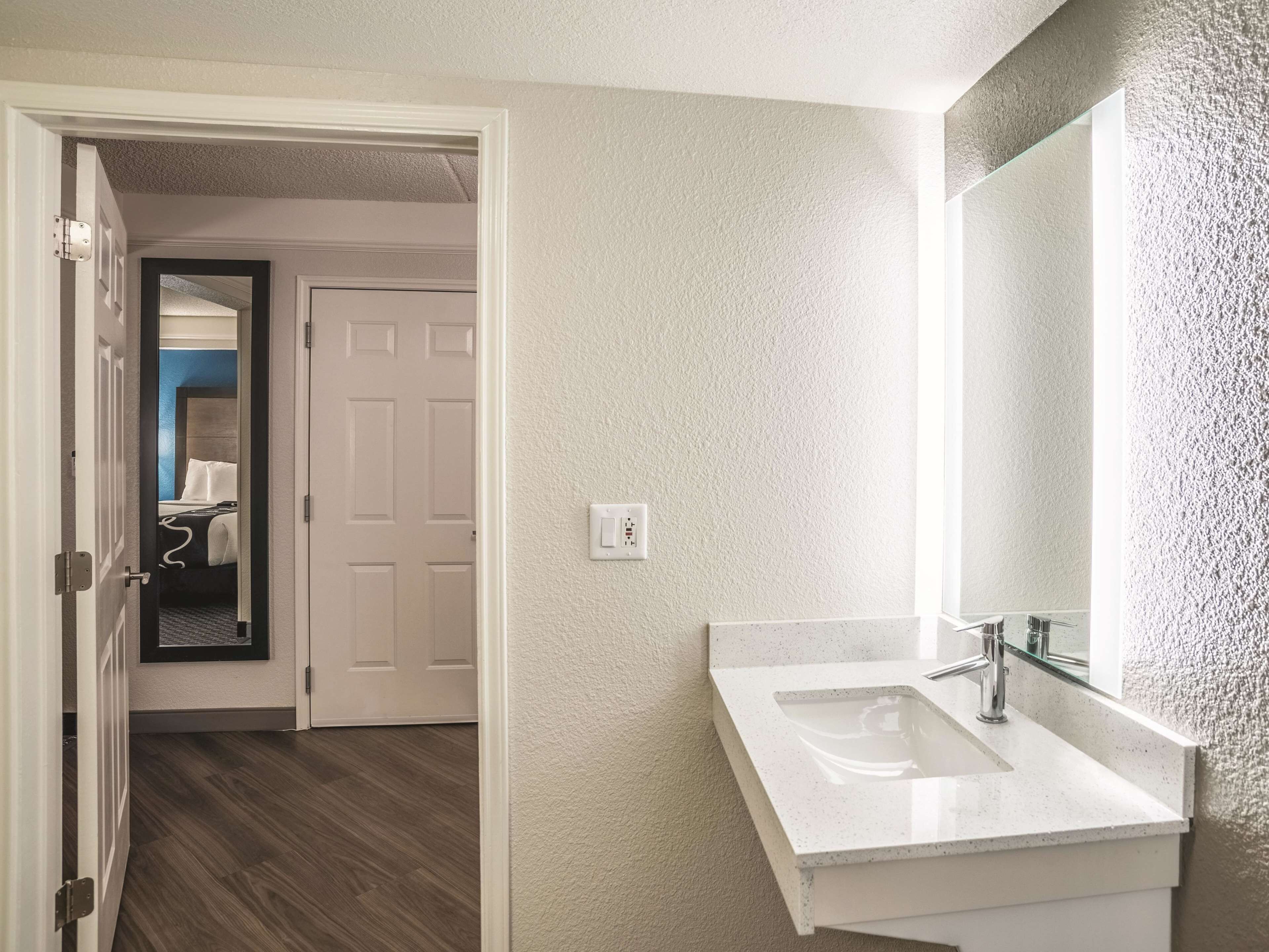 A hotel bathroom with a white sink, a large mirror, and a door leading to a bedroom.