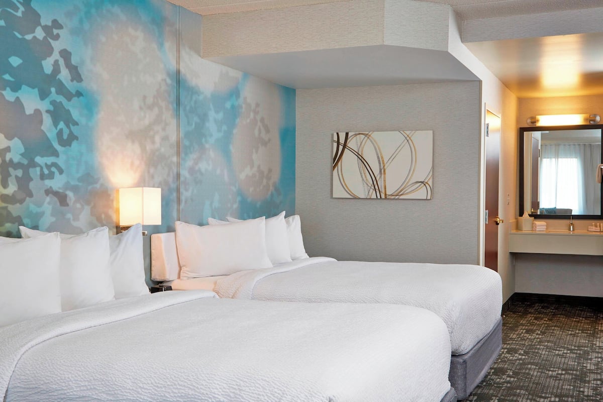 A hotel room with two beds, a large abstract painting on the wall, a mirror above the bed, and a carpeted floor.