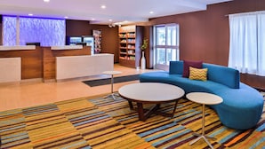 Un hall d’hôtel comprenant un comptoir d’accueil, un canapé bleu, deux tables rondes, une moquette rayée aux couleurs vives et une bibliothèque contenant divers objets.