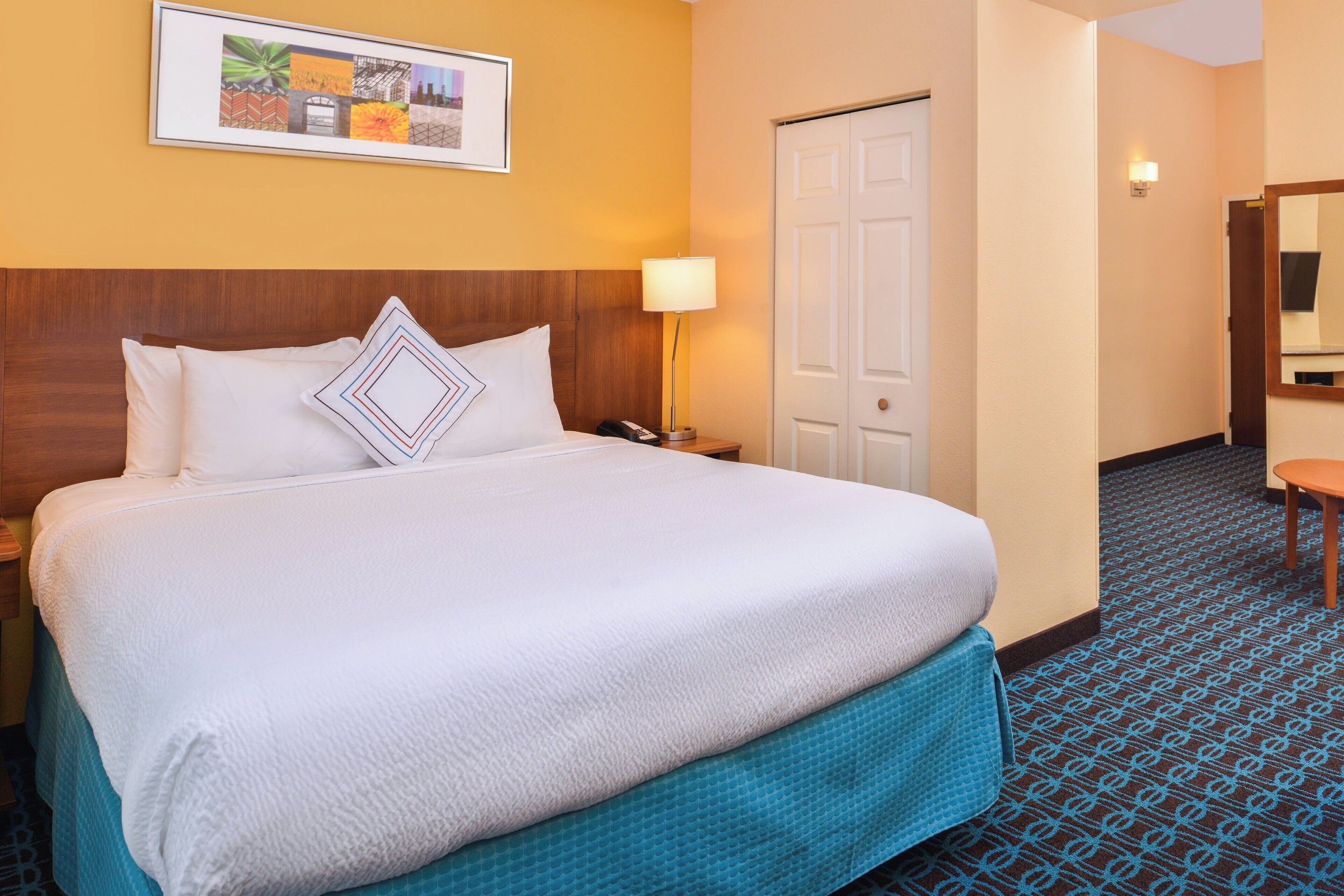 Habitación de hotel con una cama grande, una mesita de noche con lámpara, una puerta y un cuadro en la pared.