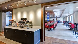 A modern hotel lobby with a long reception desk, a coffee station, and a dining area with red chairs and tables.