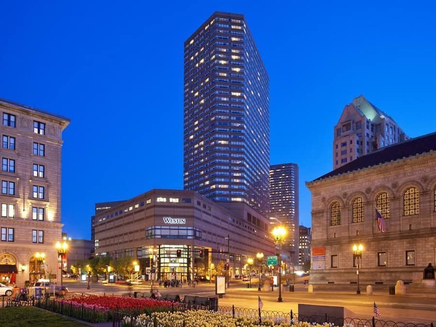 A Westin hotel with a modern facade, located in a city square with a historic building in the background.