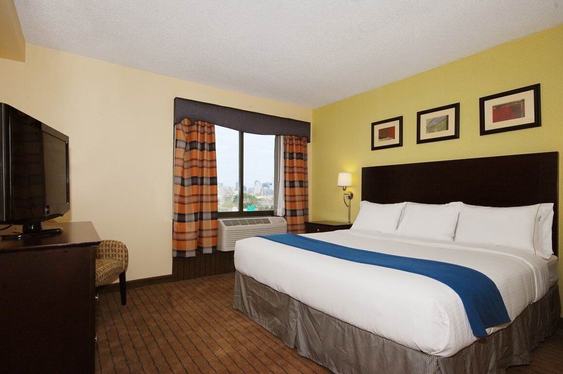 A hotel room with a bed, a television, an air conditioning unit, and a window with curtains.