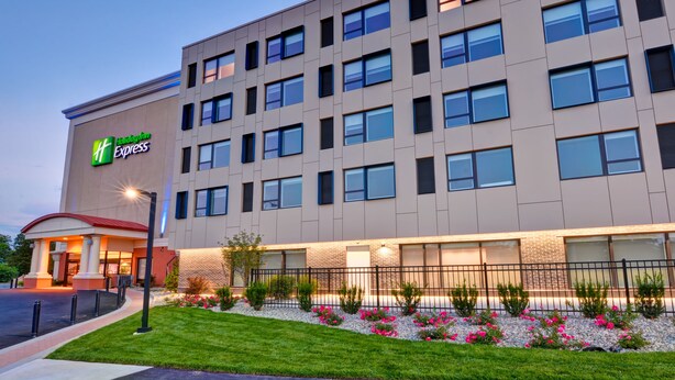 A modern hotel building with a prominent 'Holiday Inn Express' sign, multiple windows, and a well-maintained garden with flowers.