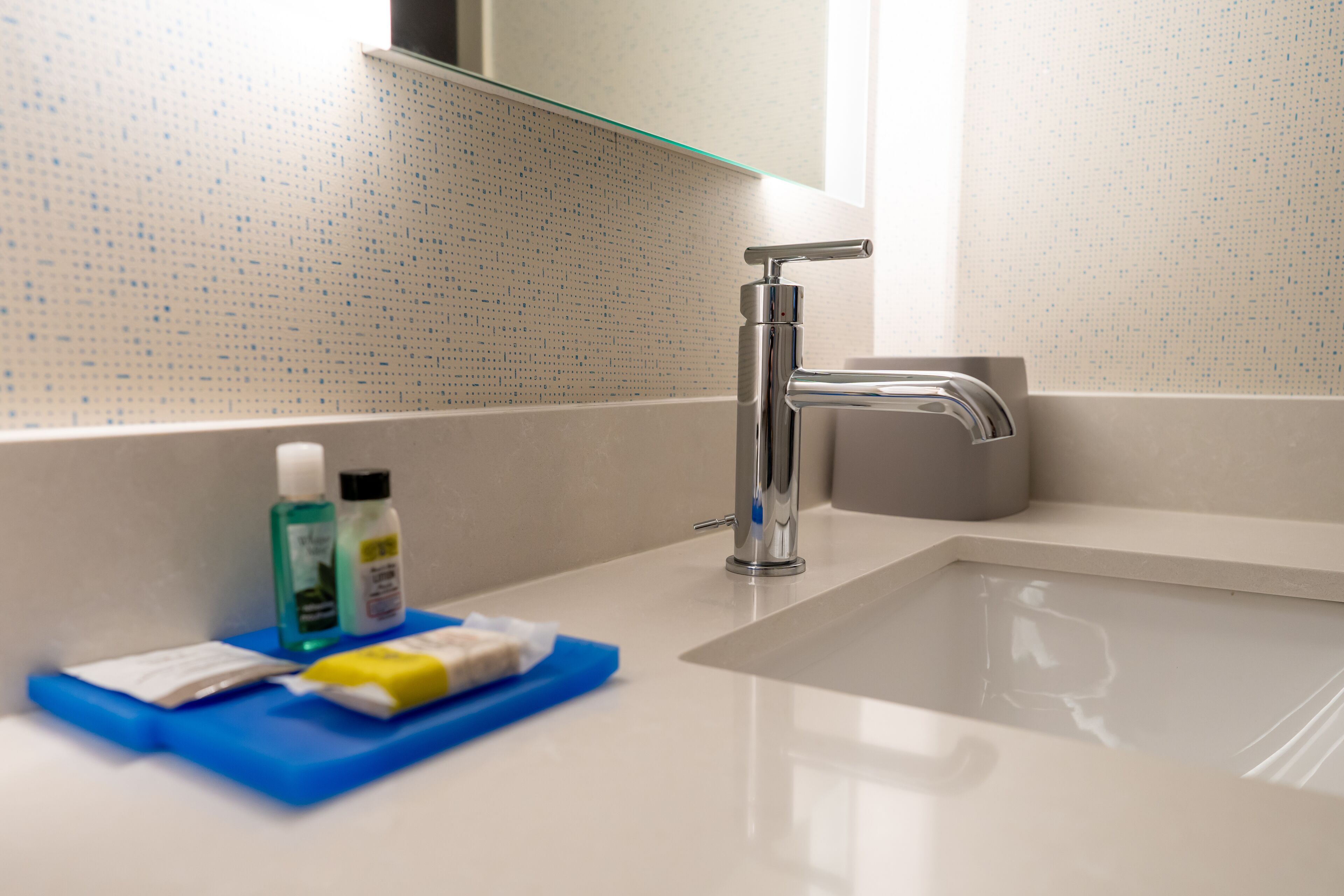 A modern bathroom with a chrome faucet, a glass shelf above the sink, and toiletries on a blue tray.