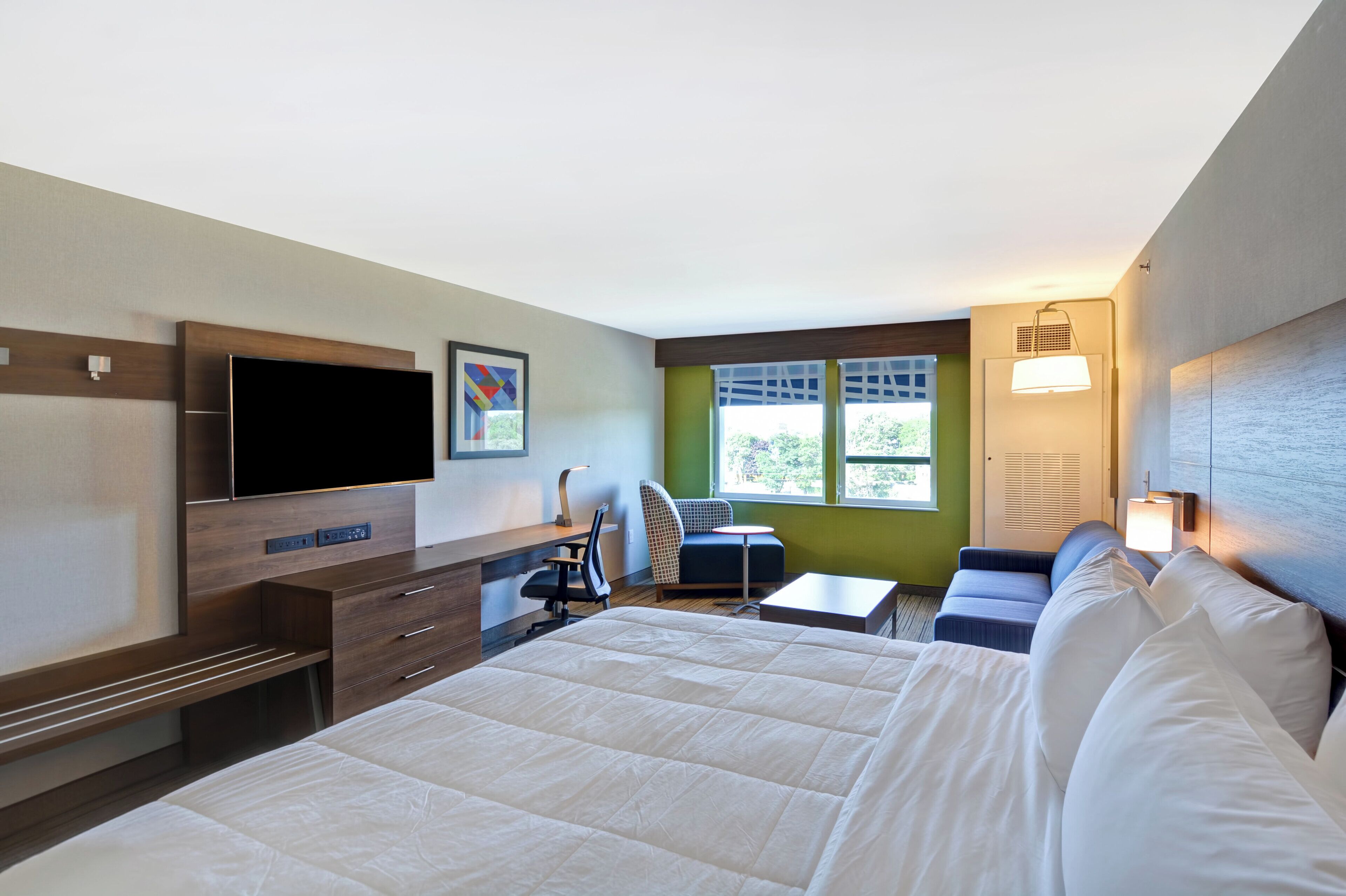 A hotel room with a bed, a desk, a chair, a TV, and a window with a view of greenery.