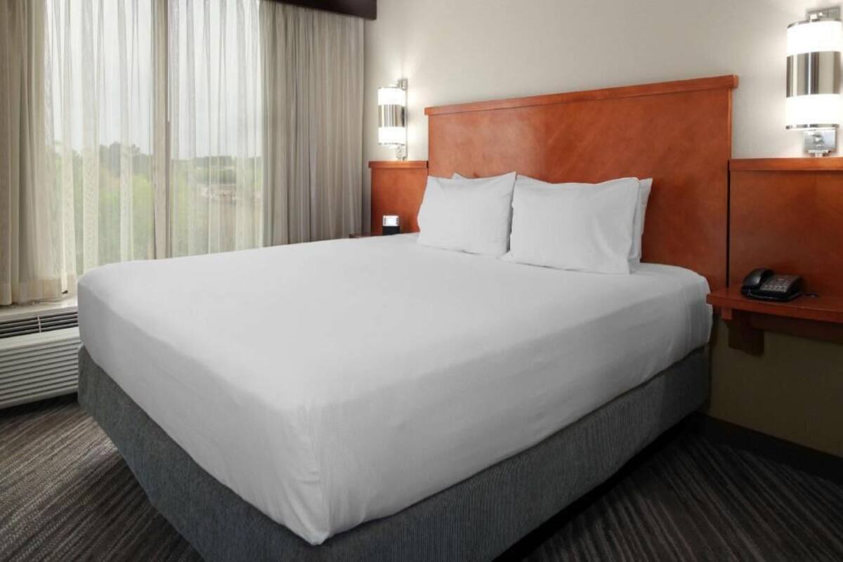 Une chambre d’hôtel avec un grand lit, une table de chevet avec un téléphone, une lampe fixée au mur et une fenêtre avec des rideaux.
