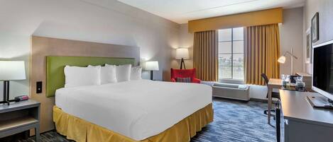Quarto de hotel com uma cama grande, uma mesa com computador, uma cadeira, televisão e janela com cortinas.