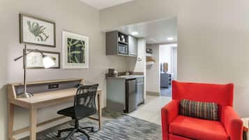 Un espacio habitable compacto con un escritorio de madera, una silla de oficina negra y un sillón rojo. Se ve una kitchenette al fondo.