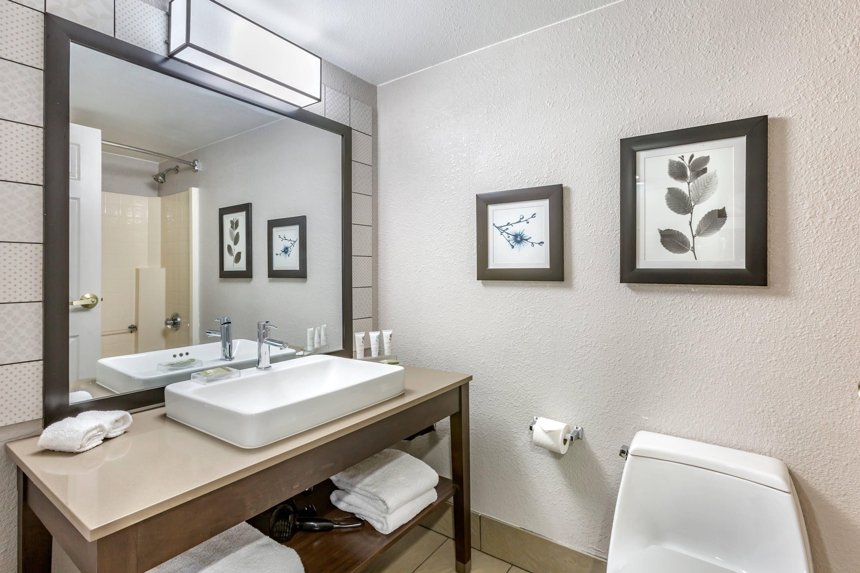 Un baño con un espejo grande, dos lavamanos, un inodoro y cuadros en la pared.