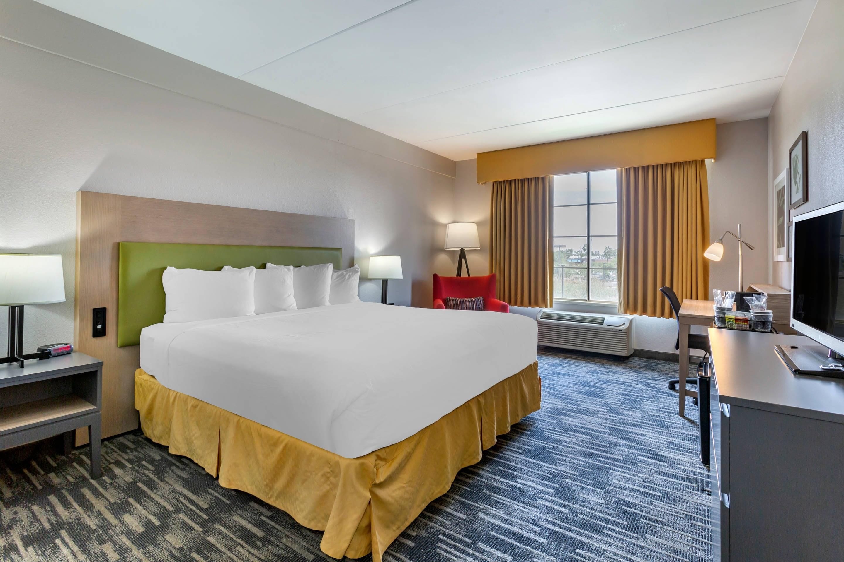 Habitación de hotel con una cama grande, un escritorio con computadora, una silla, una lámpara y una ventana con cortinas.