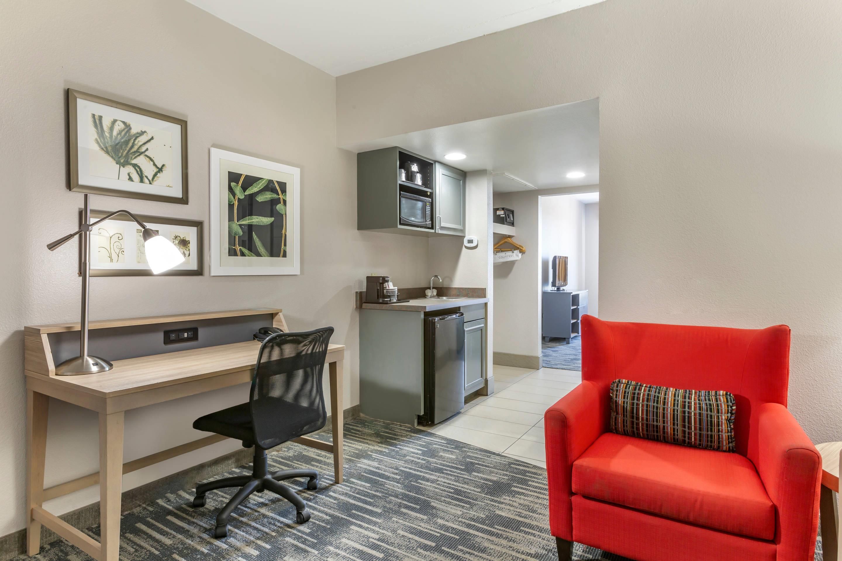 Una habitación de hotel compacta con escritorio, silla, kitchenette y un sillón rojo.