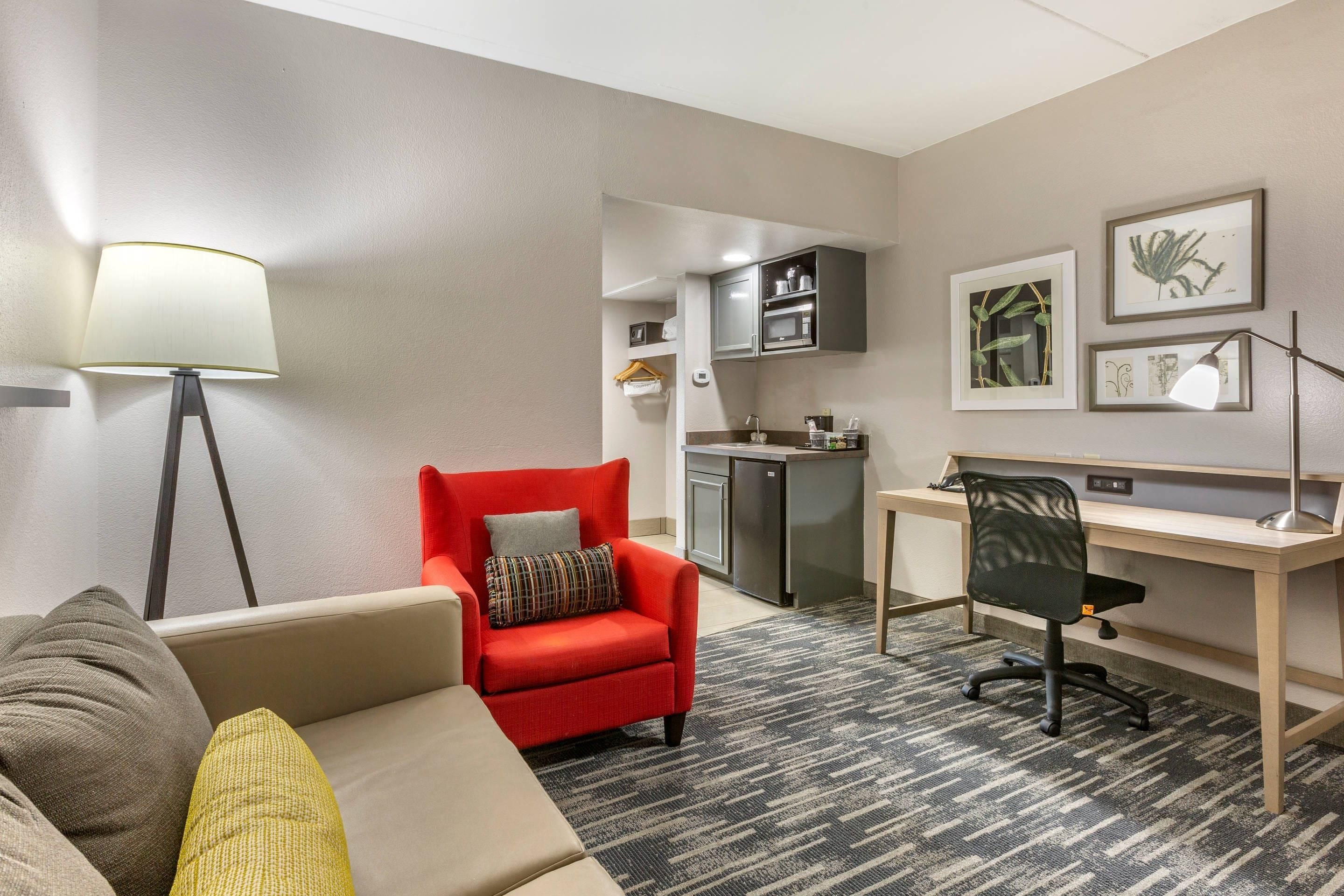 Habitación de hotel compacta con sofá, sillón rojo, escritorio con silla, kitchenette y lámpara de pie.