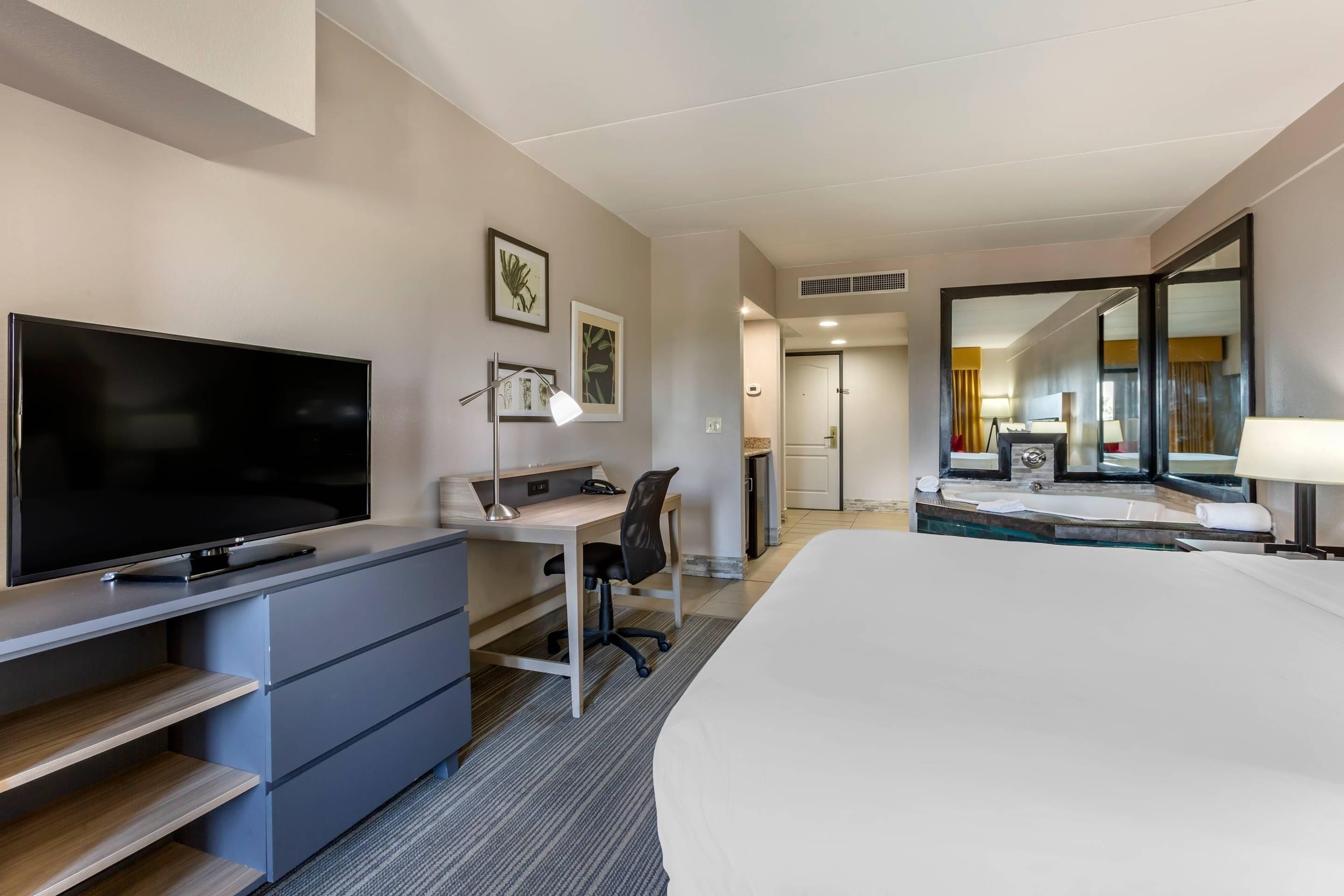 Habitación de hotel con televisión de pantalla plana, escritorio, silla, cama, baño con espejo y ventana.