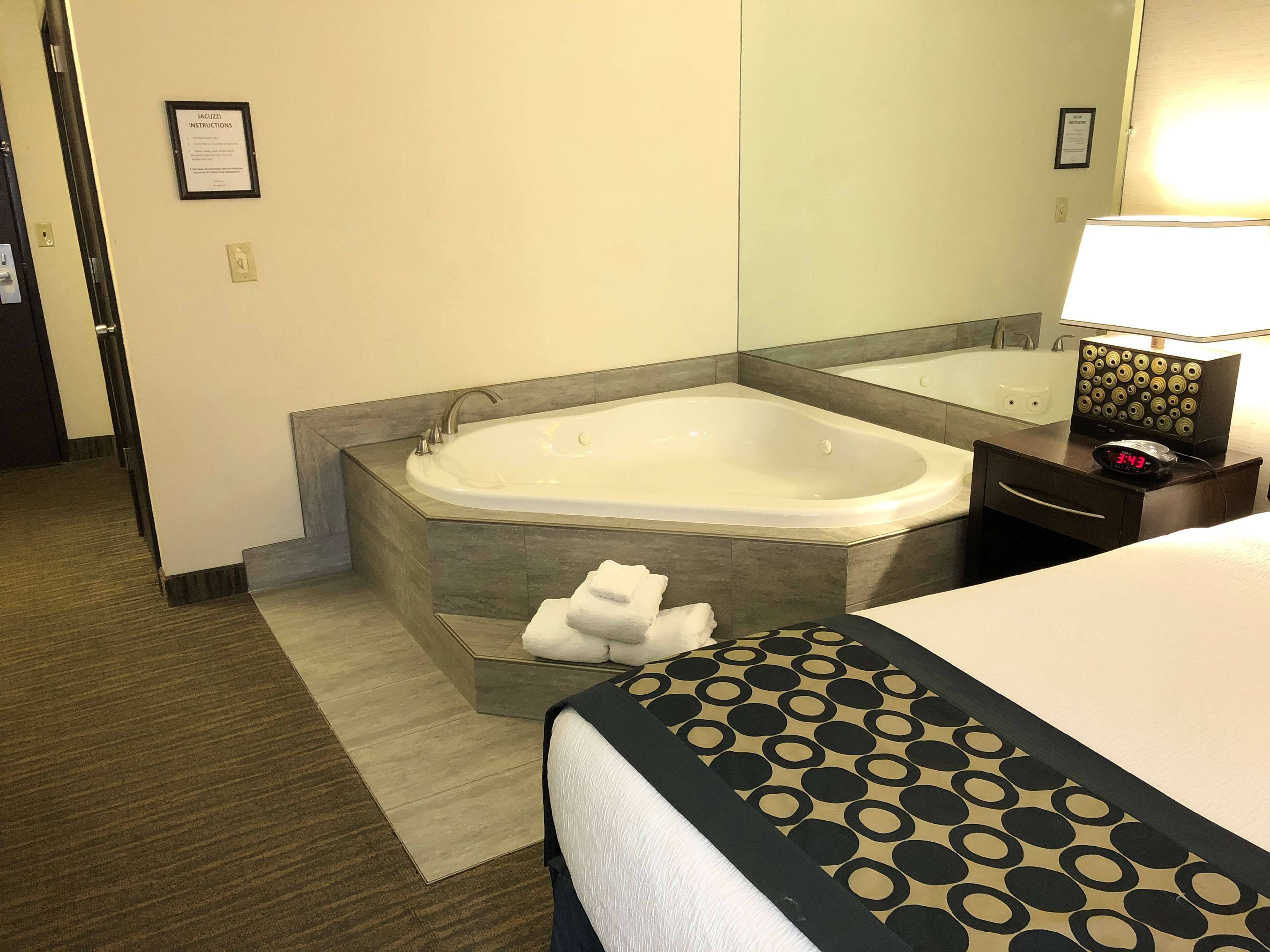 Habitación de hotel con una bañera grande, una mesita de noche con lámpara y una cama bien hecha.