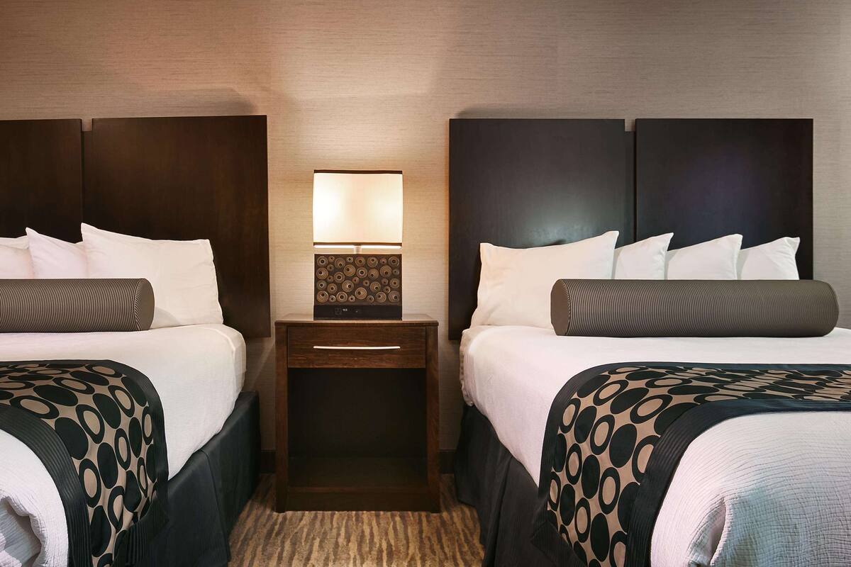Habitación de hotel con dos camas, una mesita de noche con lámpara y cabecera con diseño circular.