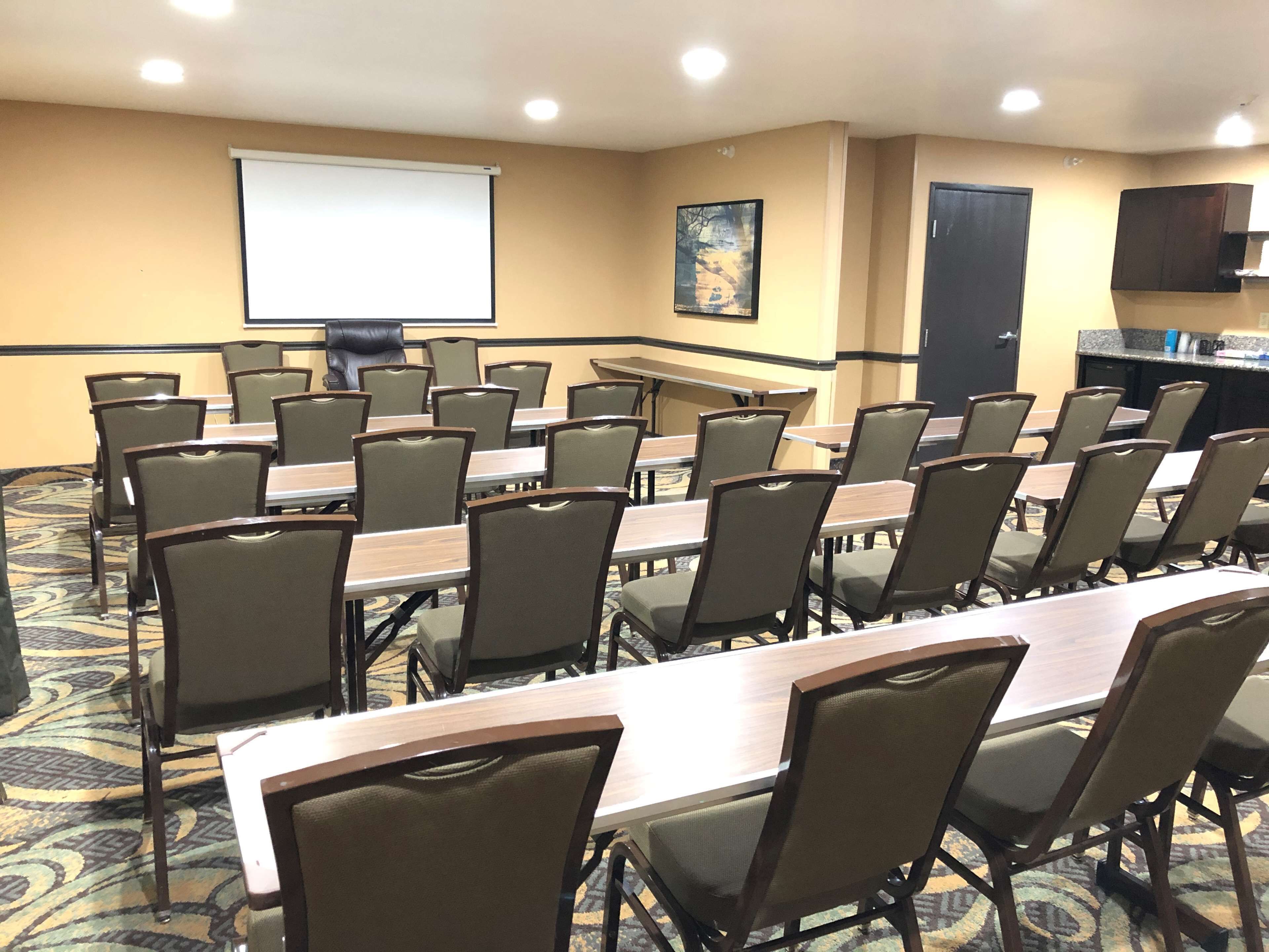 A conference room with multiple tables and chairs, a projector screen, a painting on the wall, and a kitchen area.
