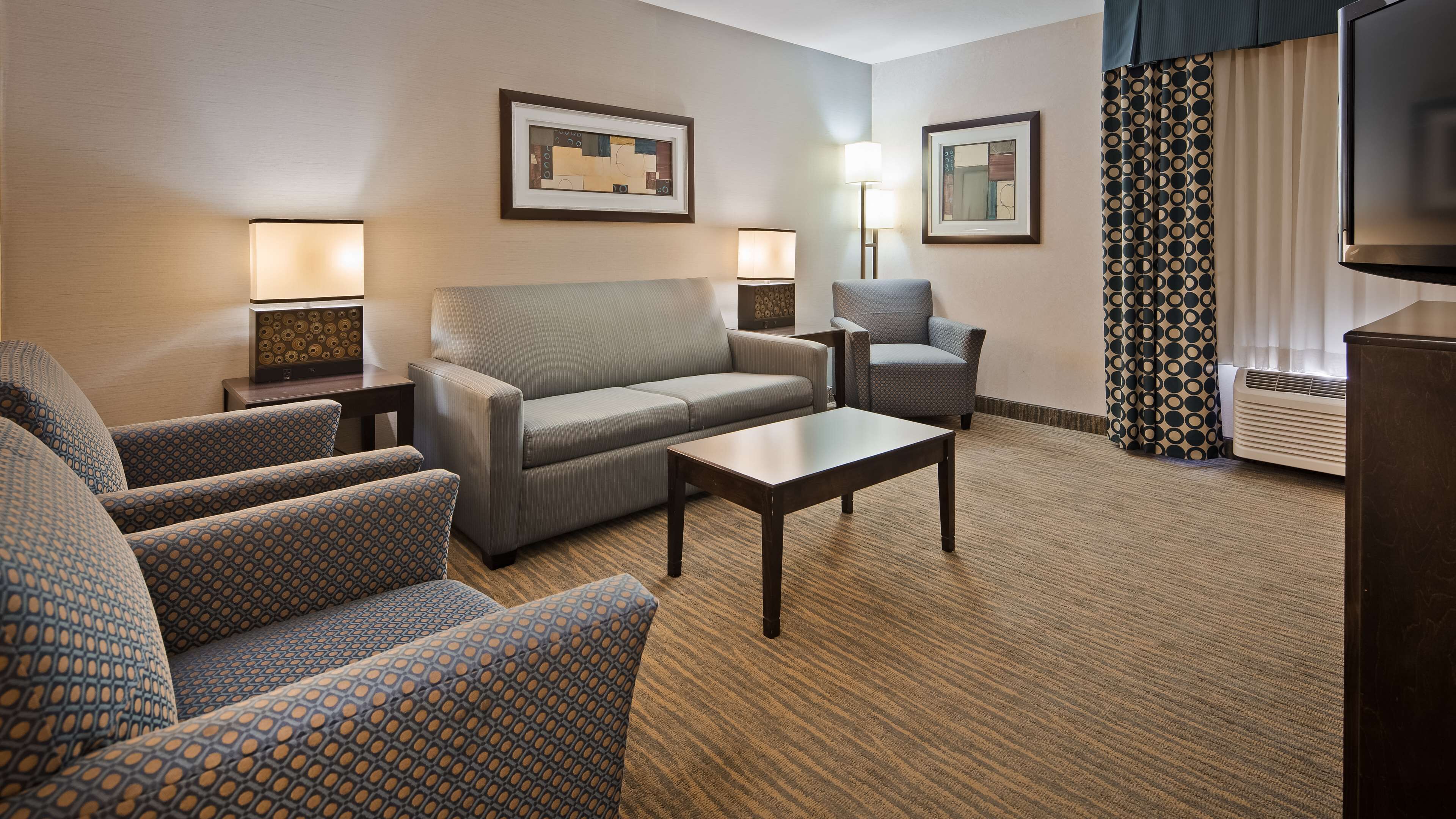 A hotel room with a sofa, armchairs, a coffee table, two lamps, and a television.