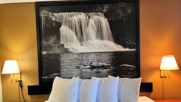 A hotel room with a large waterfall painting on the wall, a bed with white and green bedding, and two bedside lamps.