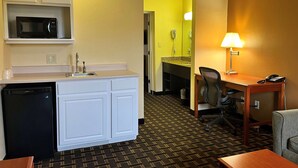 A compact hotel room with a kitchenette, a desk with a lamp, and a chair.