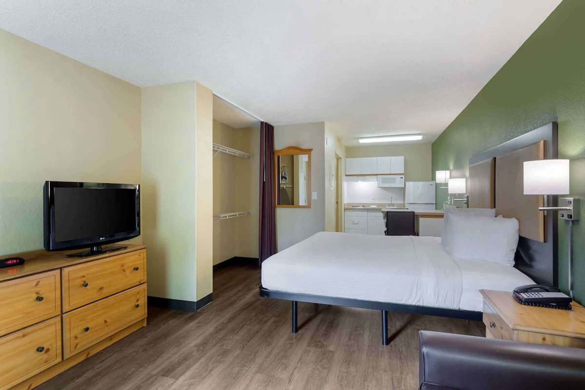 A hotel room with a bed, a TV on a wooden dresser, a desk with a lamp, and a bathroom visible through an open door.