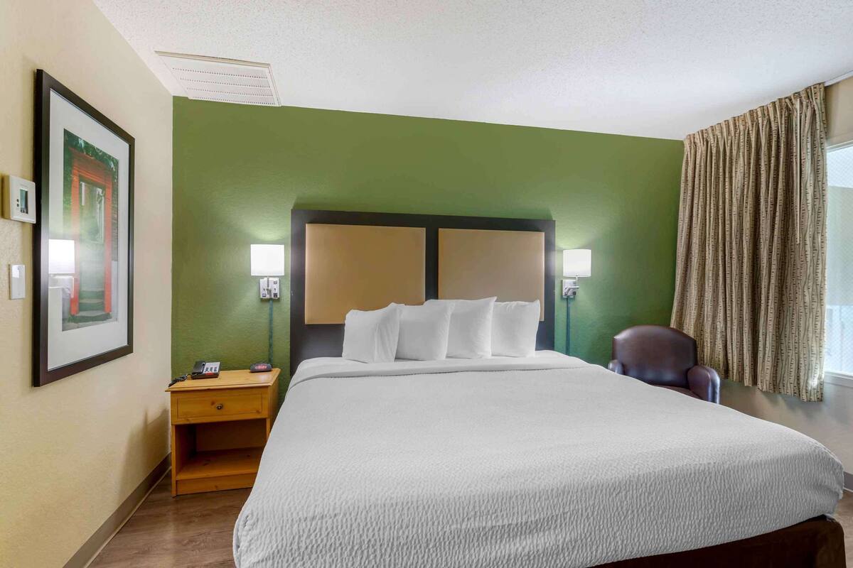 A hotel room with a large bed, two bedside lamps, a nightstand, a chair, and a window with curtains.
