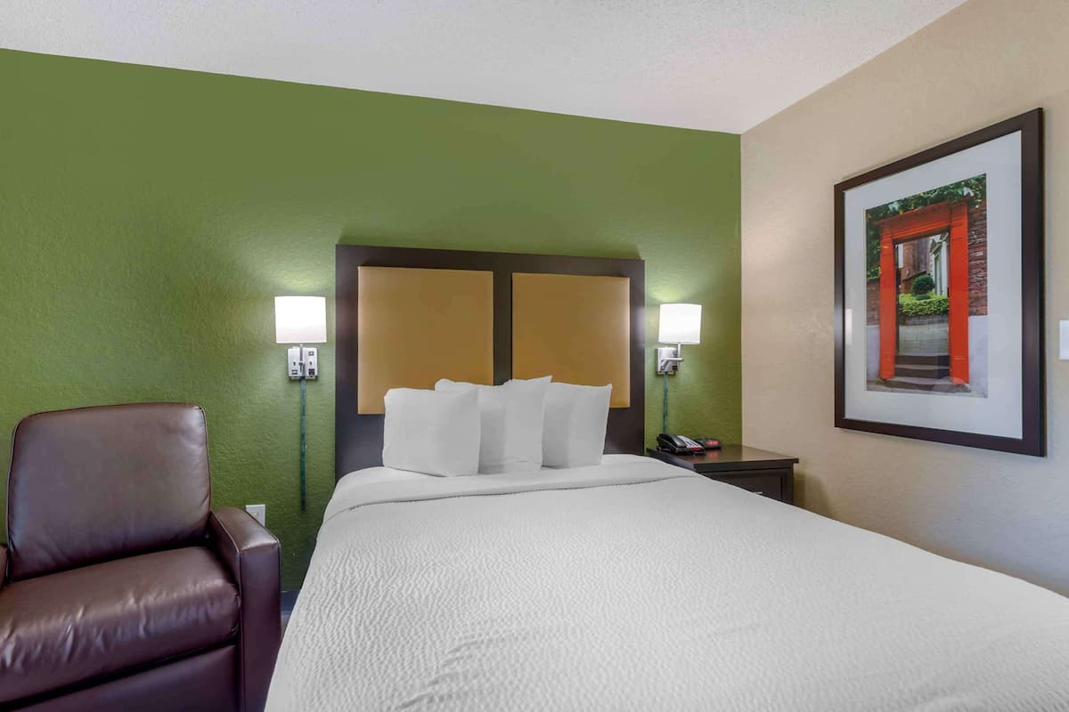 A hotel room with a bed, a brown leather chair, two bedside lamps, and a framed picture on the wall.