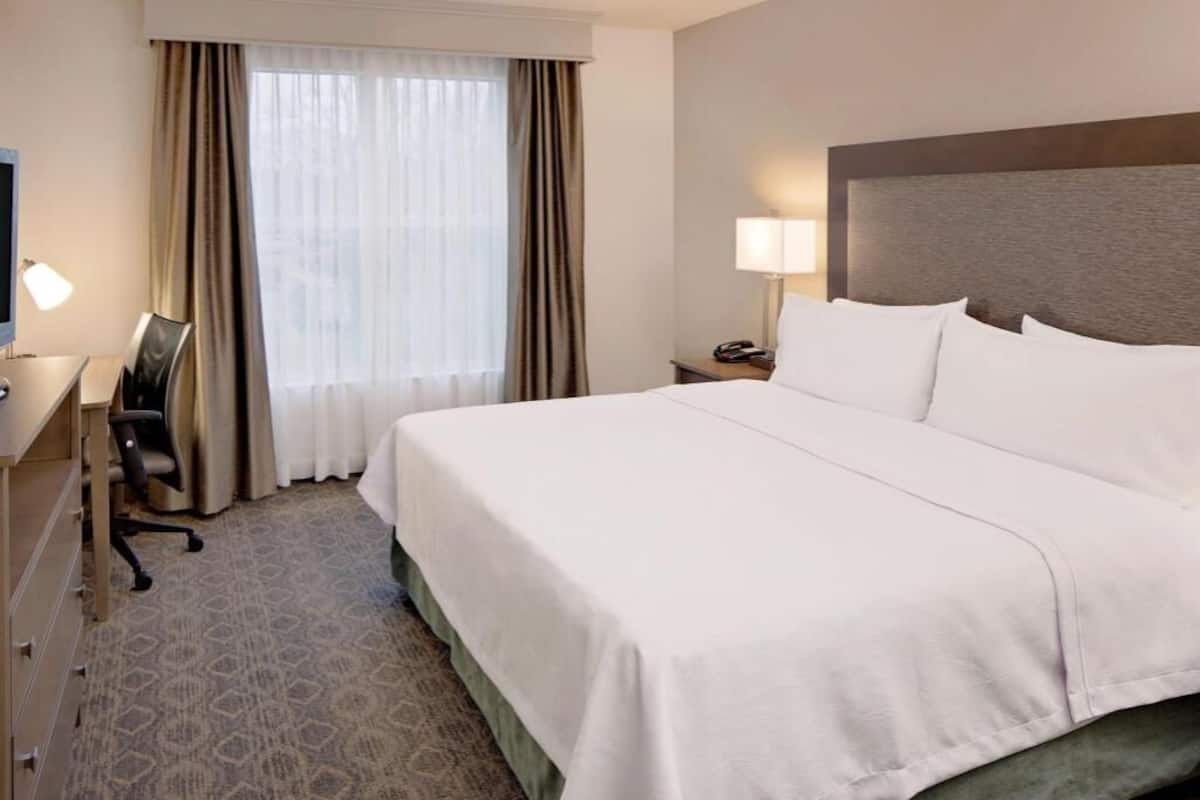 Ein Hotelzimmer mit einem großen Bett, einem Schreibtisch mit Stuhl, einer Lampe und einem Fenster mit Vorhängen.