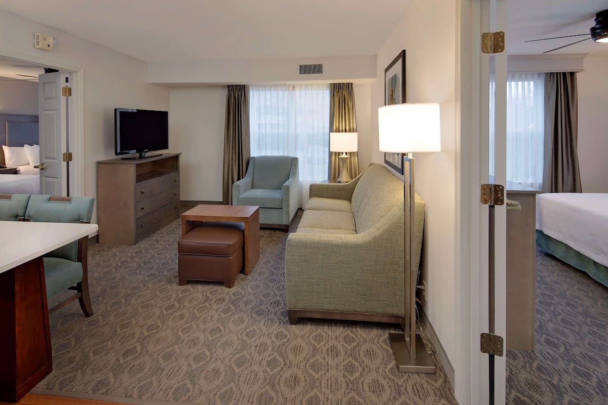 Ein Hotelzimmer mit Bett, Schreibtisch, Fernseher, Sofa, Sessel, Lampe und Tür.