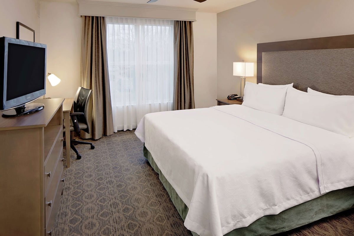 Ein Hotelzimmer mit Bett, Schreibtisch mit Fernseher, Sessel, Nachttisch mit Lampe und einem Fenster mit Vorhängen.