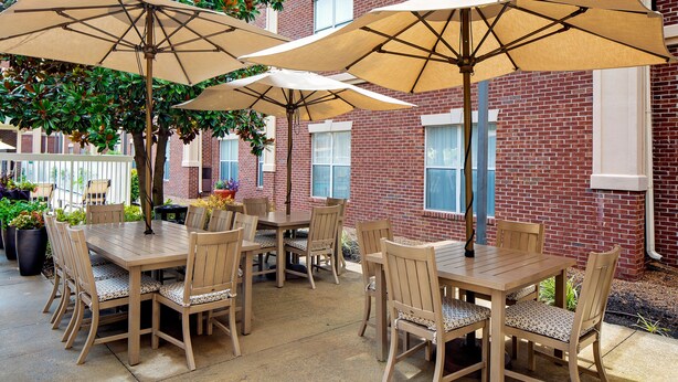 An outdoor patio with wooden tables and chairs, beige umbrellas, and a brick building in the background. - Homewood Suites by Hilton Nashville-Brentwood (Brentwood)