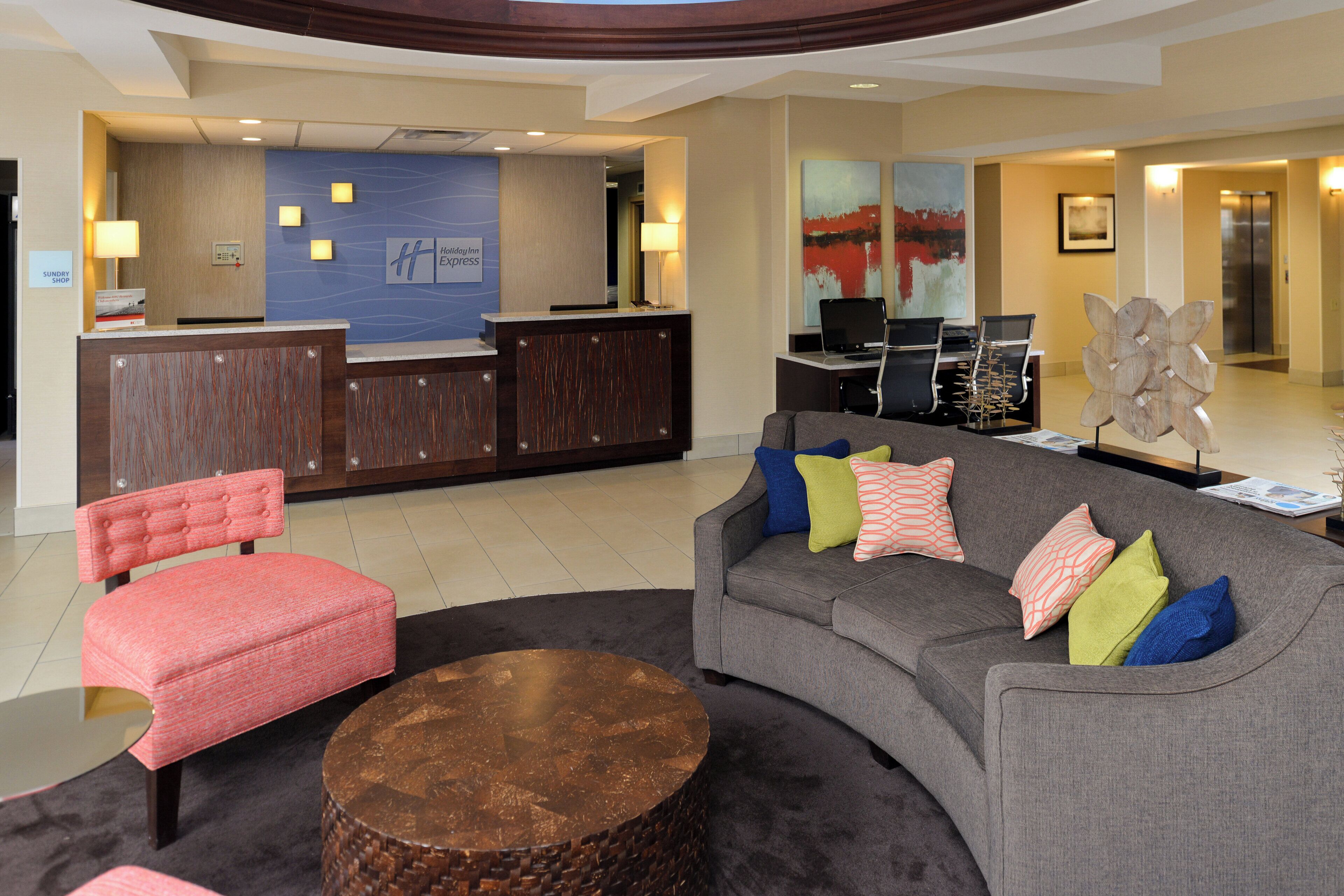 A hotel lobby with a reception desk, a grey sofa with colorful cushions, a red upholstered chair, and a round wooden coffee table.
