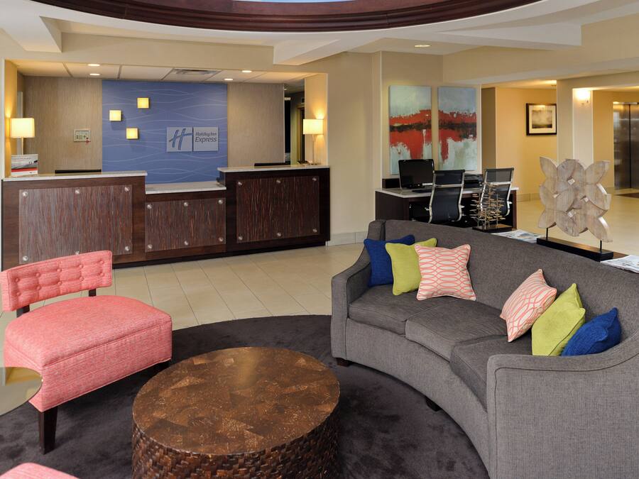 A hotel lobby with a reception desk, a grey sofa with colorful cushions, a red upholstered chair, and a round wooden coffee table.