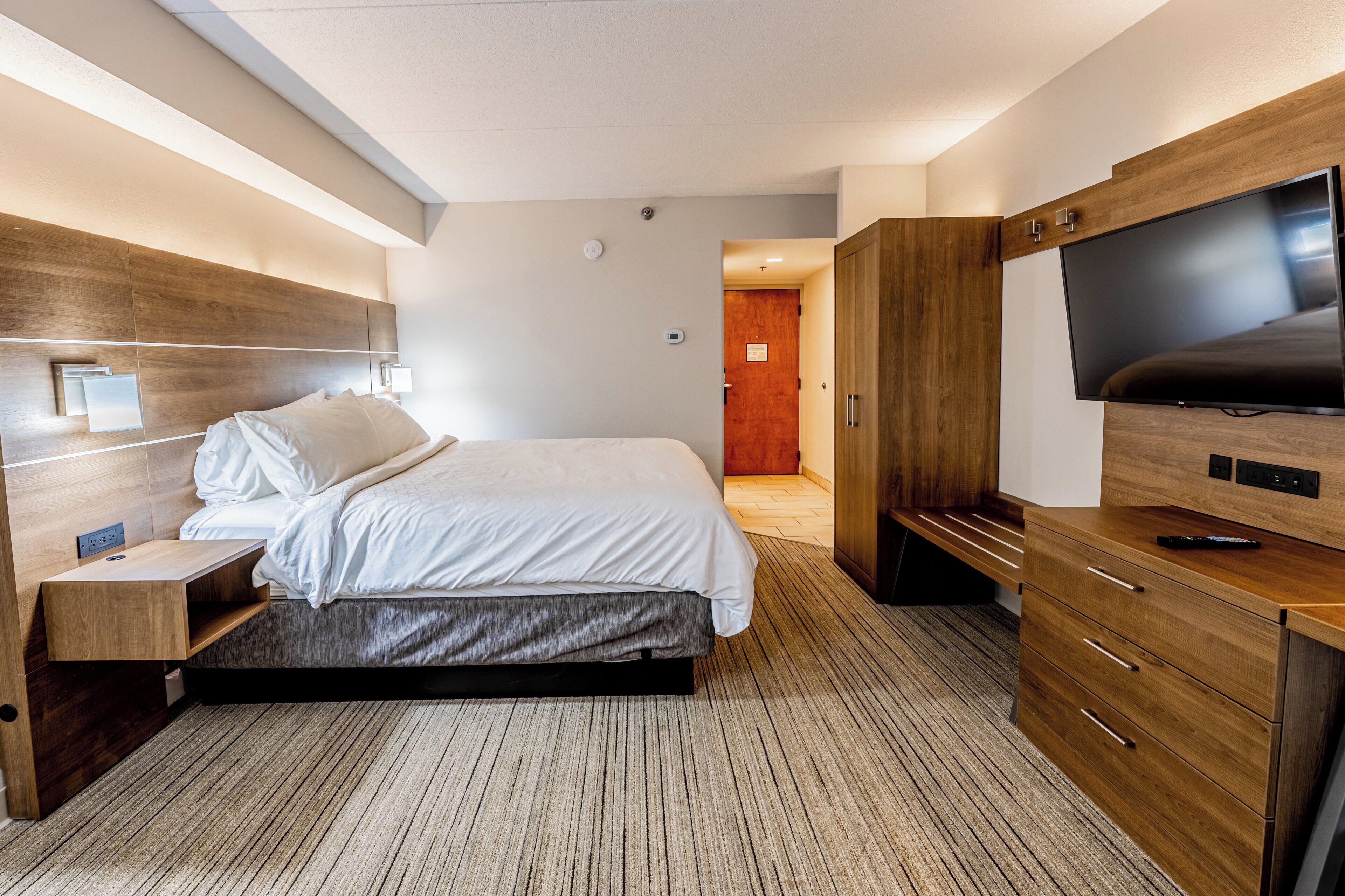 A modern hotel room with a bed, bedside table, TV, wooden wardrobe, and a carpeted floor.
