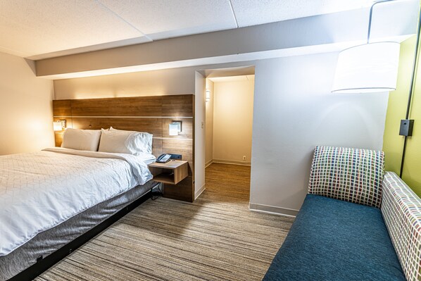 Una habitación de hotel con una cama, una mesita de noche y un pequeño rincón de descanso con un cojín estampado.