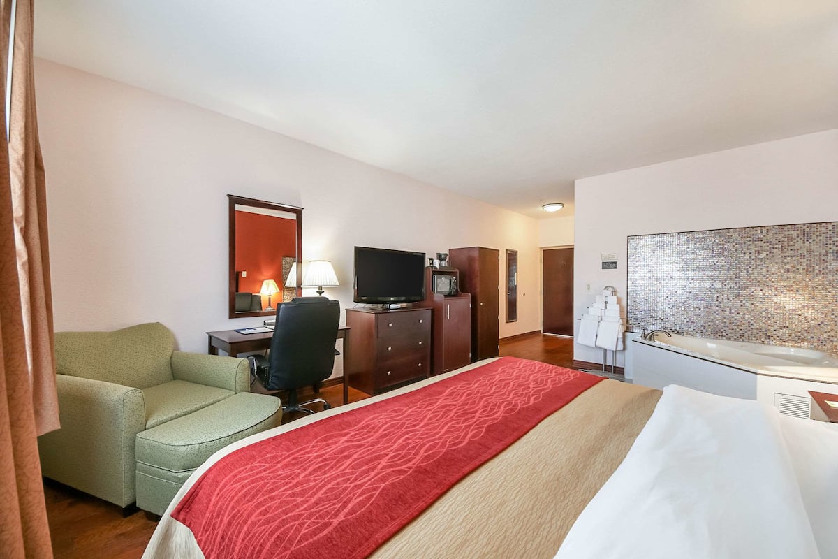 A hotel room with a bed, a desk with a television, a chair, a desk lamp, a mirror, a bathroom with a sink and mirror, and a decorative wall.