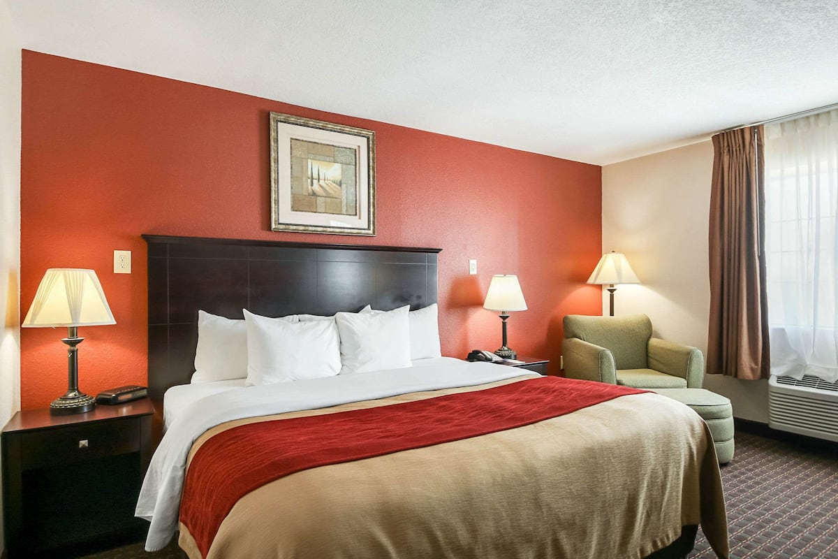 A hotel room with a large bed, two bedside tables with lamps, a chair, a framed picture on the wall, and a window with curtains.