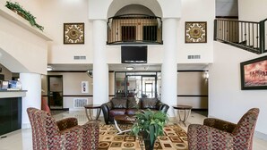 A spacious lobby with a high ceiling, a central seating area with patterned chairs, a decorative plant, and a staircase with a balcony.