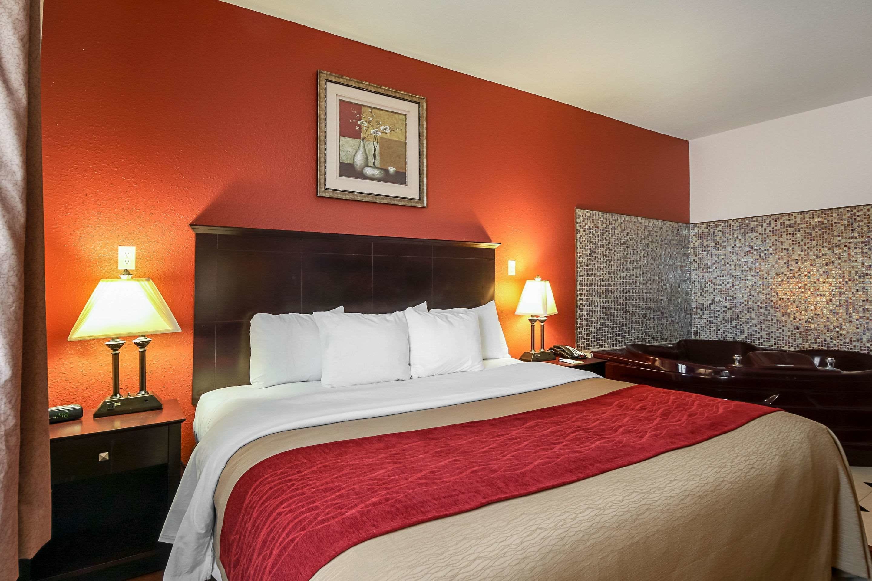 A hotel room with a large bed, two bedside lamps, a painting on the wall, a red throw blanket, and a jacuzzi tub.
