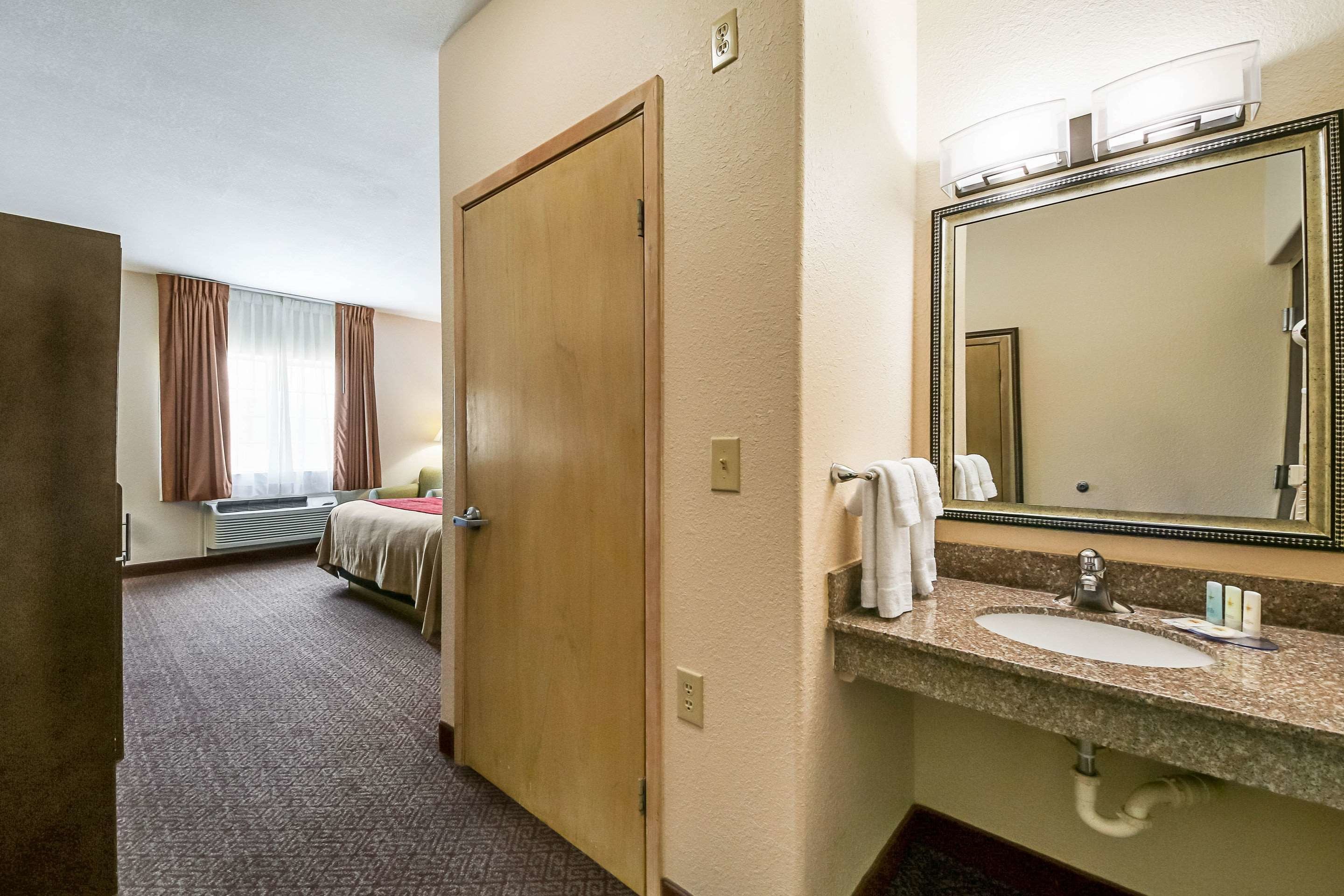 Habitación de hotel con cama, baño con lavamanos y espejo, y una puerta que da a otra habitación.